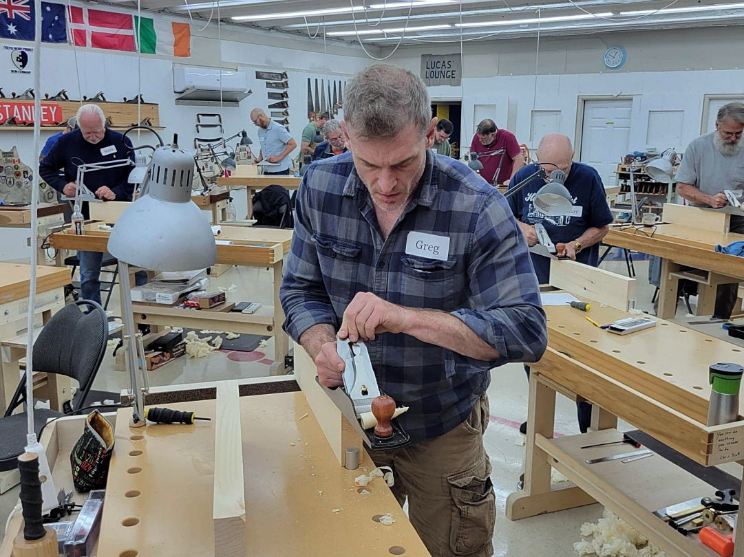 Man working with a tool in a workshop setting with other people and equipment around.