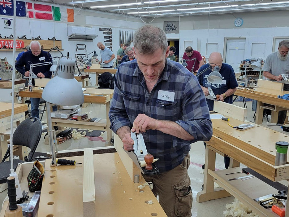 Man working with a tool in a workshop setting with other people and equipment around.
