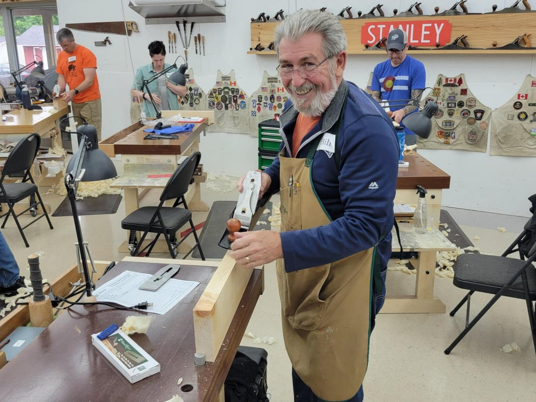 Man in a workshop using a Stanley tool, surrounded by woodworking equipment and other people.