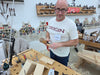A man wearing glasses and a white t-shirt with 'ORIGIN' printed on the chest stands in a woodworking workshop, holding a wooden chisel. The background features a workbench, tools, and wooden panels.