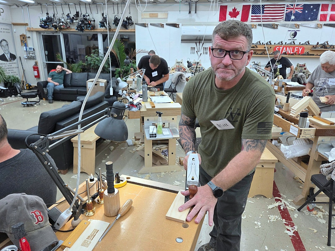 A man in a woodworking workshop holding a white and silver metal sliding block tool, surrounded by workbenches, tools, and other people working in the background.