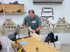Man working at a wooden workbench with tools and aprons in the background