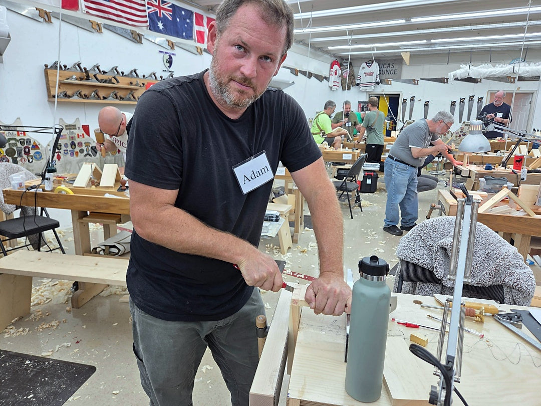 A man wearing a black t-shirt and gray pants works at a woodworking table inside a workshop, using a chisel on a piece of wood. A gray water bottle is on the table next to him, and other people and woodworking tools are visible in the background.