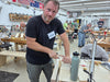 A man wearing a black t-shirt and gray pants works at a woodworking table inside a workshop, using a chisel on a piece of wood. A gray water bottle is on the table next to him, and other people and woodworking tools are visible in the background.