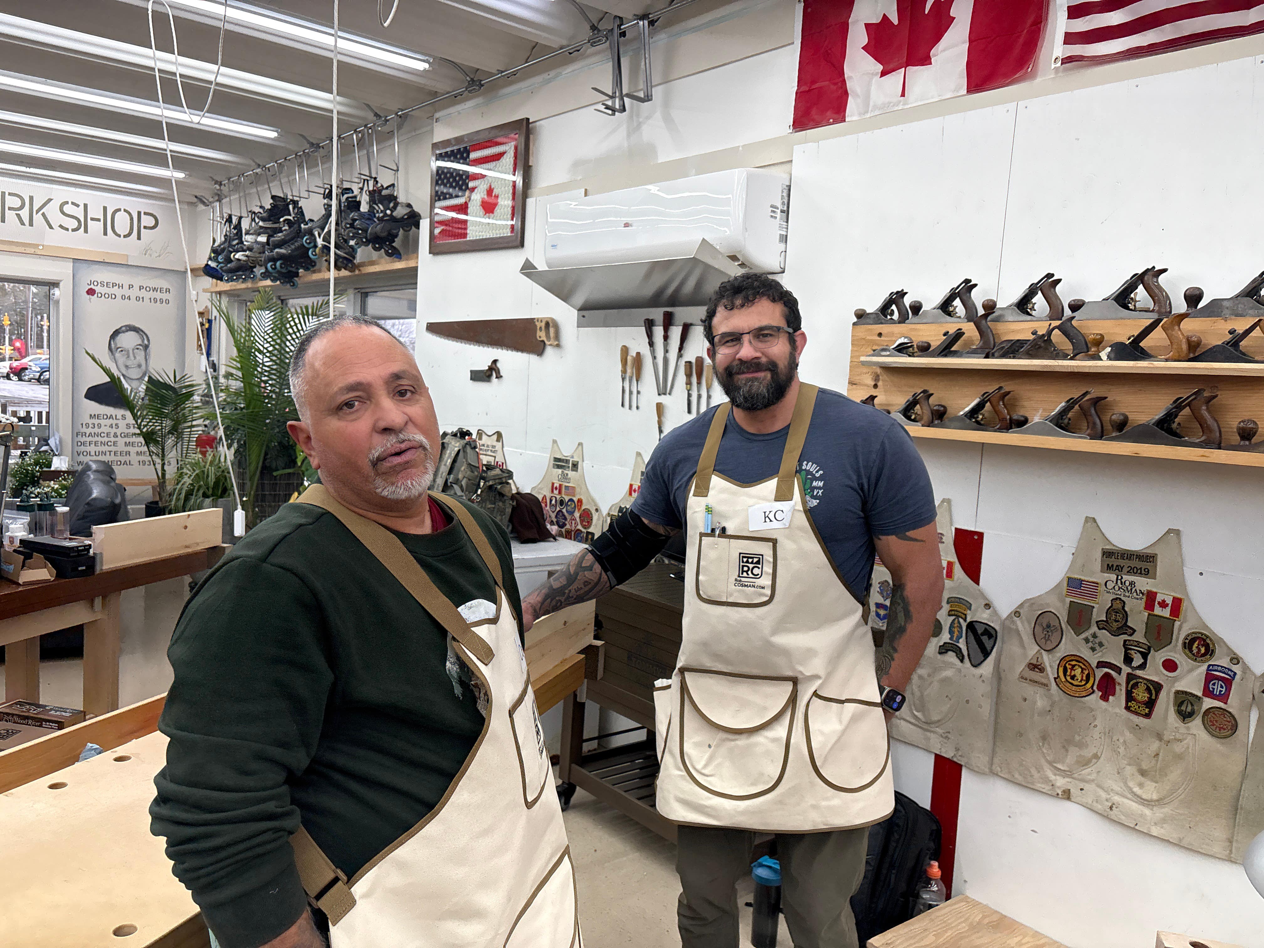 Two men in a workshop wearing aprons, with tools and equipment in the background.