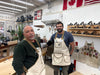 Two men in a workshop wearing aprons, with tools and equipment in the background.