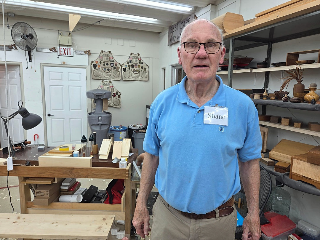 An older man with glasses and a name tag reading 'Shane' stands in a cluttered woodworking workshop. He is wearing a light blue polo shirt and khaki pants, surrounded by shelves of wood, tools, and workbenches.