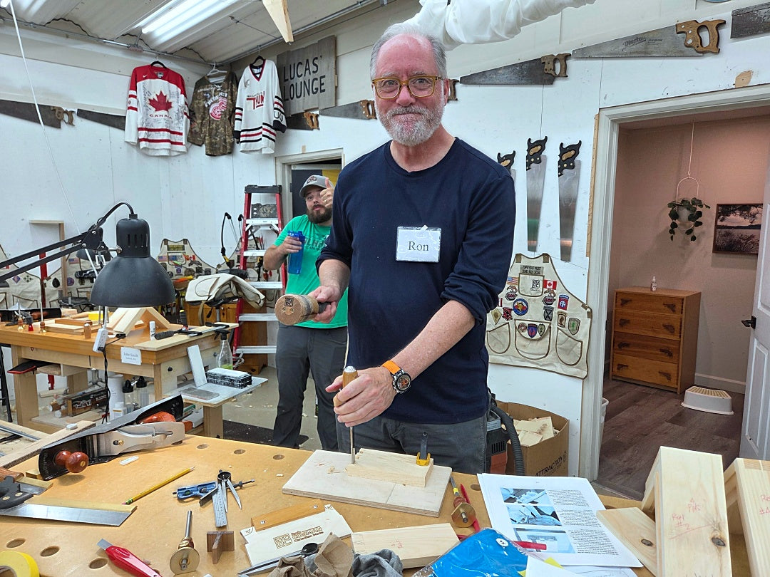A man with glasses and a beard wearing a navy blue long-sleeve shirt stands in a cluttered woodworking workshop, holding a small wooden block. Another man in a green shirt is visible in the background.