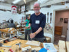 A man with glasses and a beard wearing a navy blue long-sleeve shirt stands in a cluttered woodworking workshop, holding a small wooden block. Another man in a green shirt is visible in the background.