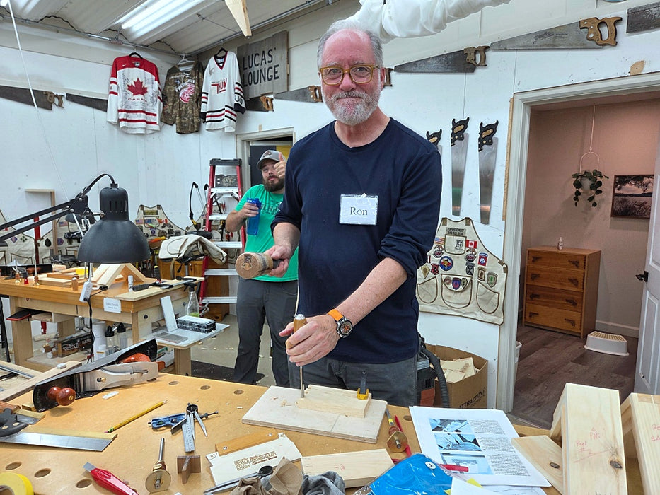 A man with glasses and a beard wearing a navy blue long-sleeve shirt stands in a cluttered woodworking workshop, holding a small wooden block. Another man in a green shirt is visible in the background.