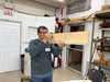 Man holding a wooden board in a workshop with 'Best Care Anywhere' sign in the background