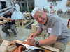 An older man with white hair smiles while working at a woodworking bench in a workshop, using a hand plane. Another person is seated at a table in the background, and a third person stands further back.