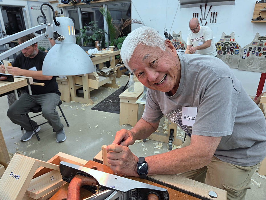 An older man with white hair smiles while working at a woodworking bench in a workshop, using a hand plane. Another person is seated at a table in the background, and a third person stands further back.