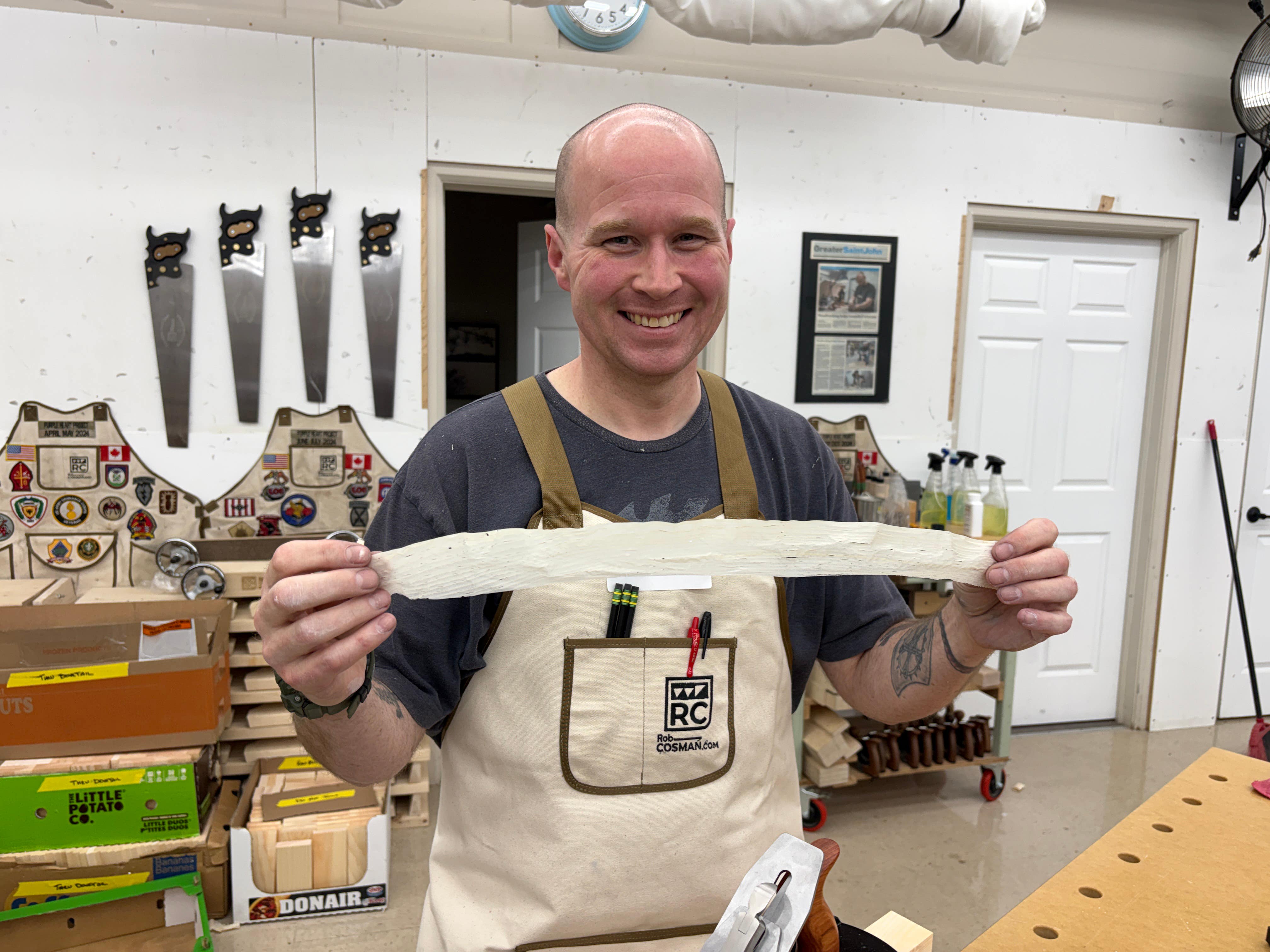 Man in a workshop holding a long white object, wearing an apron with visible branding.