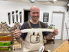 Man in a workshop holding a long white object, wearing an apron with visible branding.