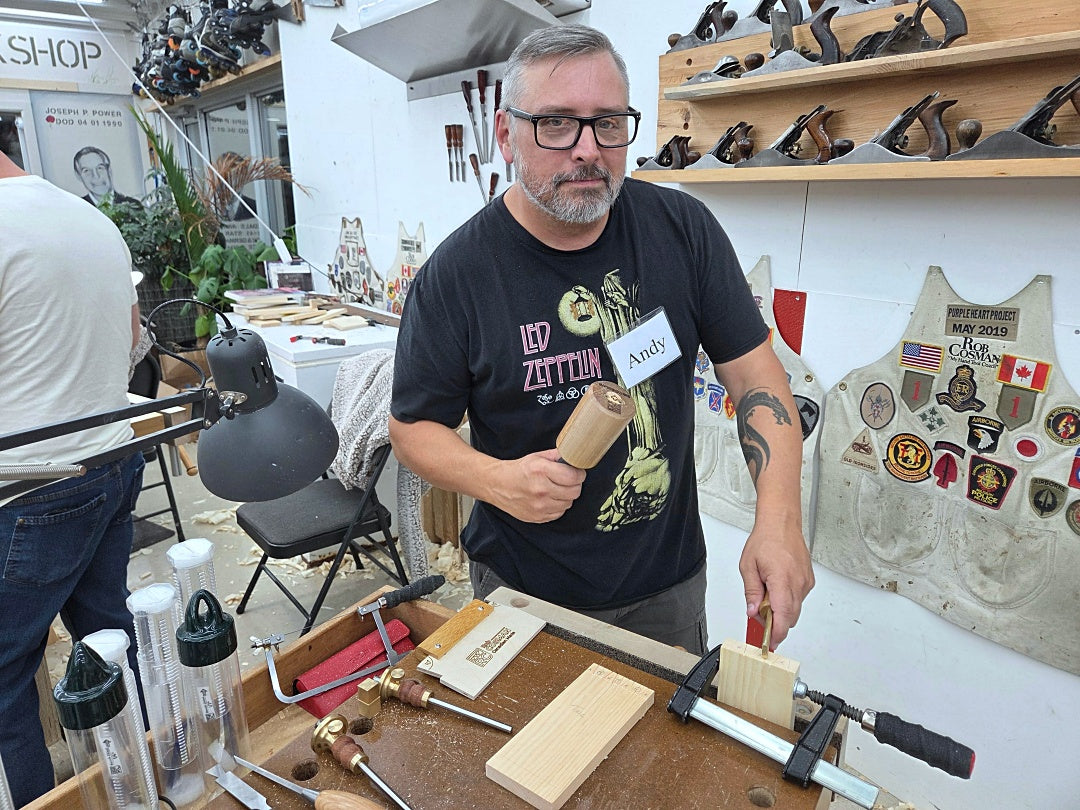 A man wearing a black graphic t-shirt with a 'Led Zeppelin' print and a name tag, holding a small wooden block and mallet in a workshop setting.