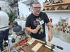 A man wearing a black graphic t-shirt with a 'Led Zeppelin' print and a name tag, holding a small wooden block and mallet in a workshop setting.