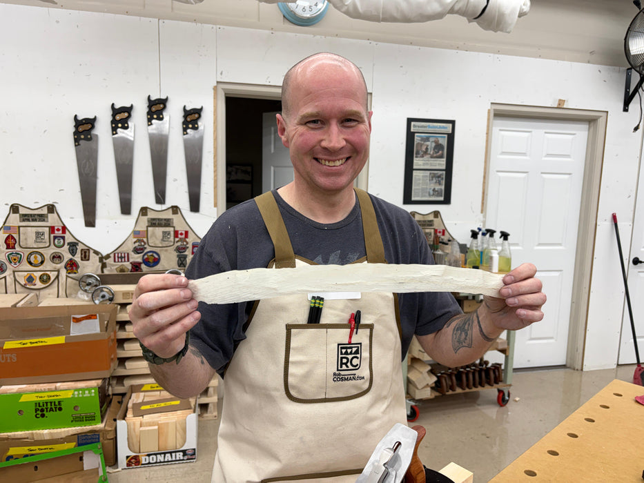 Man in a workshop holding a long white object, wearing an apron with visible branding.