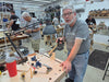 A man in a gray graphic t-shirt and jeans stands in a woodworking workshop, holding a hand saw next to a workbench with various tools. Other woodworkers are visible in the background of the shop.