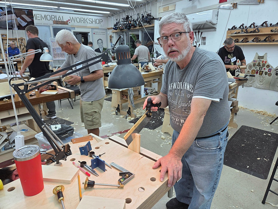 A man in a gray graphic t-shirt and jeans stands in a woodworking workshop, holding a hand saw next to a workbench with various tools. Other woodworkers are visible in the background of the shop.