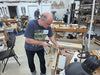 A man in a woodworking workshop wearing a navy blue graphic t-shirt and black pants, using a square and pencil at a workbench. The workshop is filled with tools, shelves, and other woodworking equipment.