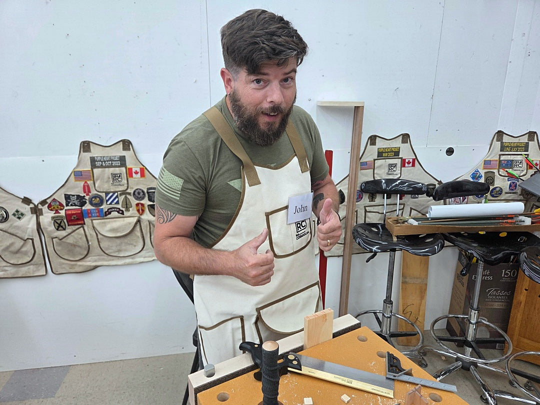 A bearded man wearing a beige canvas apron over an olive green t-shirt stands in a workshop, gesturing with two thumbs toward the camera. Other canvas aprons with patches are hanging on the wall behind him.
