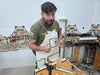 A bearded man wearing a beige canvas apron over an olive green t-shirt stands in a workshop, gesturing with two thumbs toward the camera. Other canvas aprons with patches are hanging on the wall behind him.