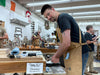Man in a workshop wearing an apron, surrounded by tools and equipment.