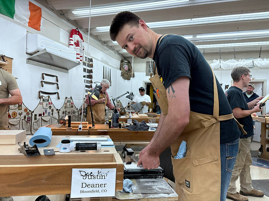Man in a workshop wearing an apron, surrounded by tools and equipment.