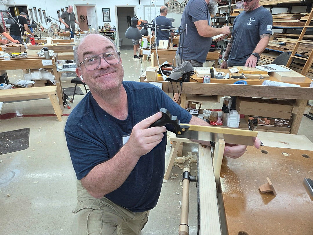 A man in a woodworking shop wearing a navy blue t-shirt and khaki pants, holding a wooden square and miter box. Other woodworkers are visible in the background of the workshop.