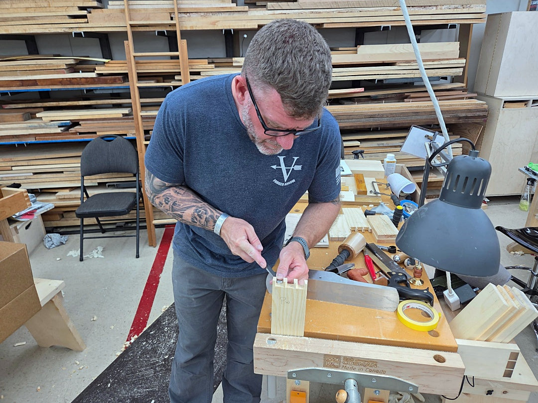 A man wearing a navy blue t-shirt and gray pants stands at a woodworking bench in a workshop, using a chisel on a piece of light-colored wood. The workshop is filled with wood stacks, tools, and a workbench lamp.