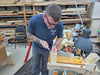 A man wearing a navy blue t-shirt and gray pants stands at a woodworking bench in a workshop, using a chisel on a piece of light-colored wood. The workshop is filled with wood stacks, tools, and a workbench lamp.