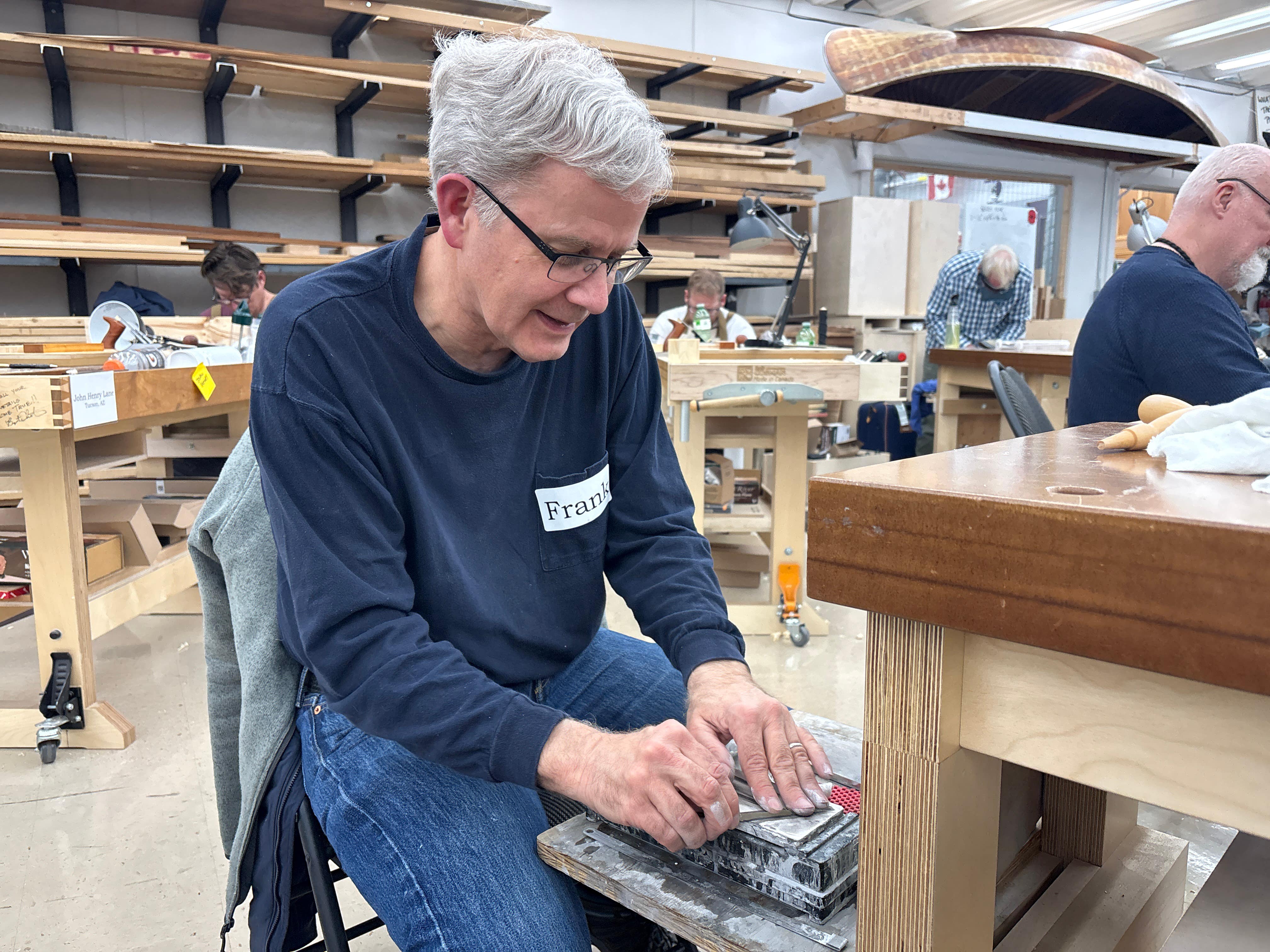 Man working on a wooden project in a workshop with other people in the background.