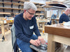 Man working on a wooden project in a workshop with other people in the background.