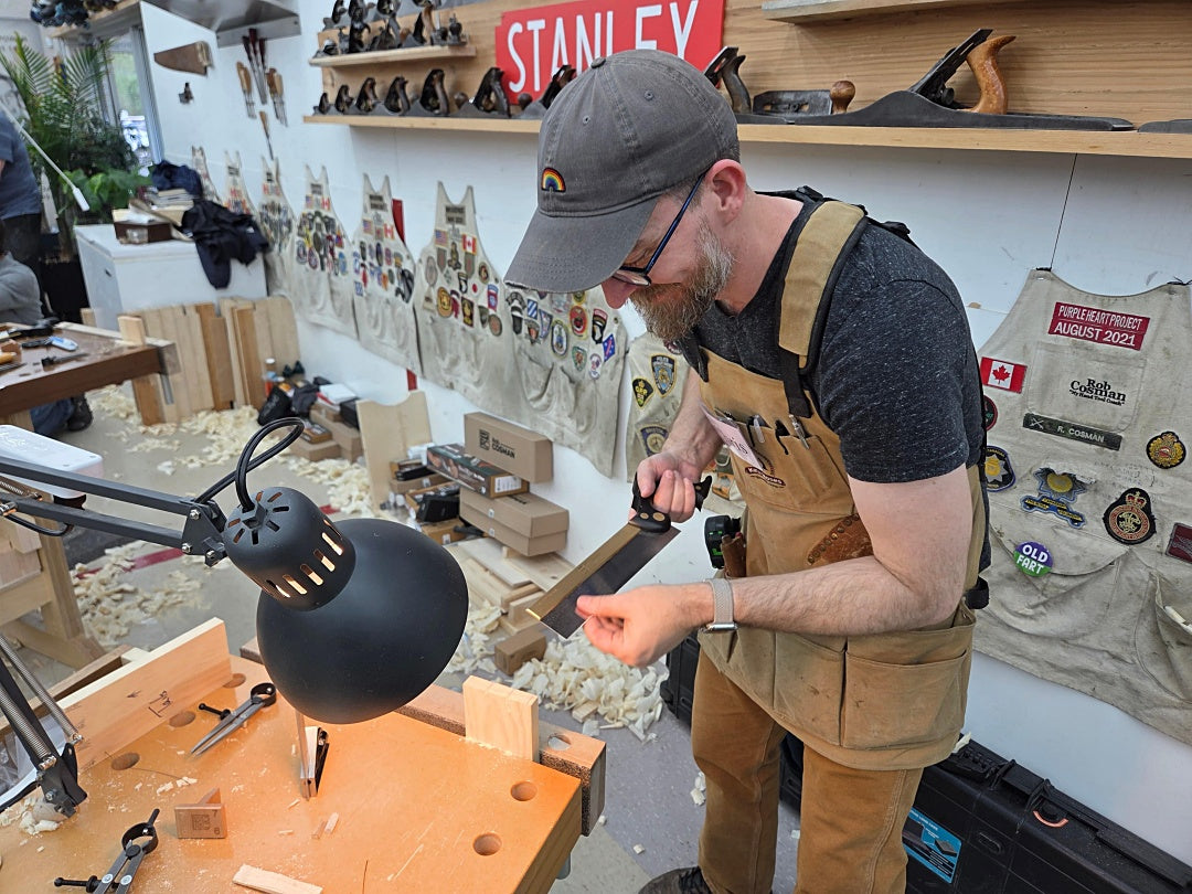 Man in a workshop wearing an apron and cap, surrounded by tools and equipment.