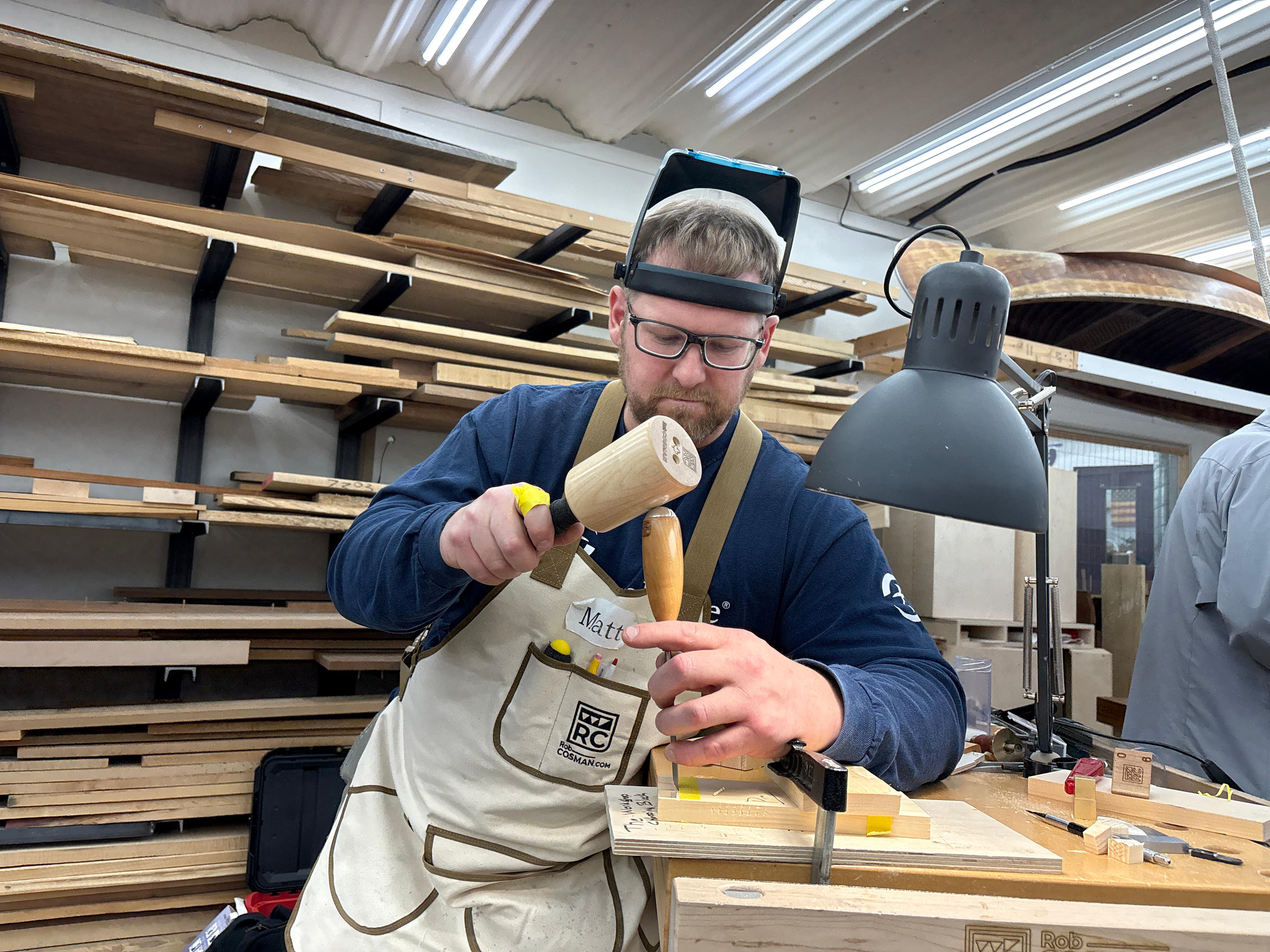 Man in a workshop using a hammer and chisel, wearing a protective helmet and apron.