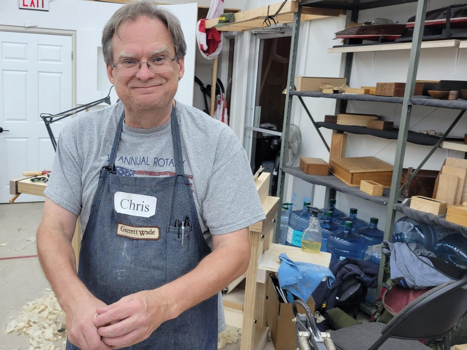 Man wearing an apron with a name tag labeled 'Chris' in a workshop setting.