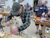 Man in a workshop wearing an apron and cap, working on a wooden project.