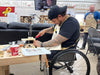 Person in a wheelchair working on a project at a table with various items, including a TV screen in the background.