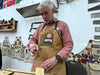 Man in a workshop wearing a brown apron with various patches, working on a wooden object.