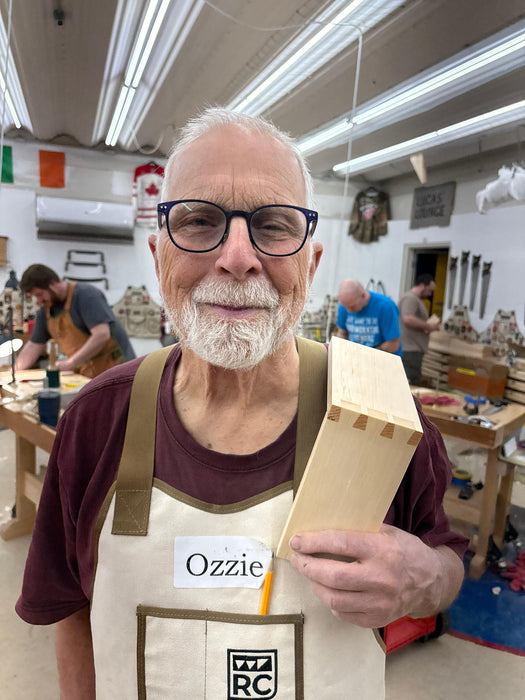 Man in a workshop holding a wooden block, wearing an apron with 'Ozzie' on it.