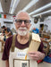 Man in a workshop holding a wooden block, wearing an apron with 'Ozzie' on it.