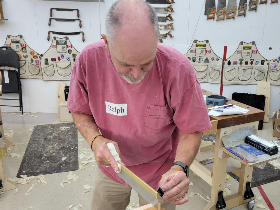 Man in a workshop wearing a pink shirt with 'Ralph' on it, working on a piece of wood.