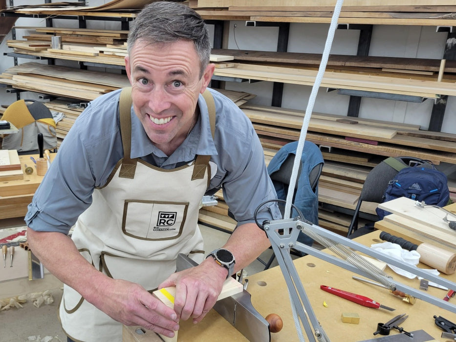 Man in a workshop wearing an apron, surrounded by wooden planks and tools.