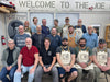 Group of people wearing aprons in a workshop setting with a sign that reads 'Welcome to The Joe'.