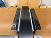A black metal woodworking vise mounting bracket on a wooden workbench, with a light blue panel visible behind them.