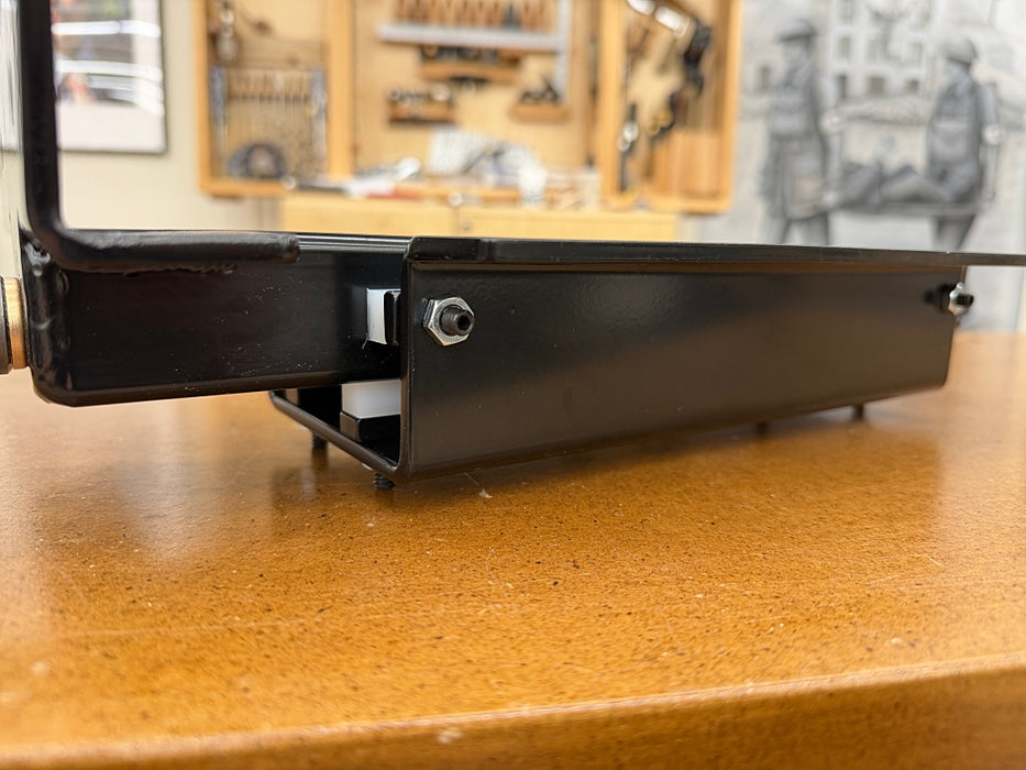 A close-up of a black metal woodworking vise bracket mounted on a wooden table, with a workshop interior blurred in the background.