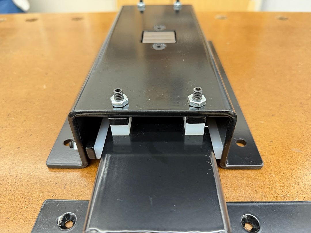 A close-up of a black metal mounting bracket or channel assembly with bolts and a sliding plate, shown on a wooden workbench.