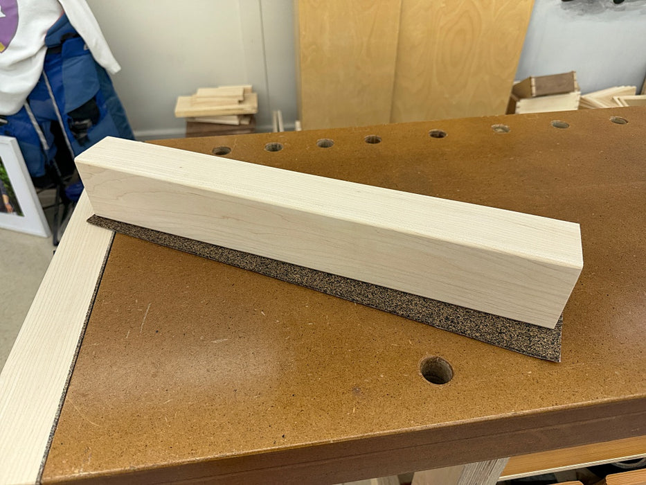 A hard maple vise jaw with a dark cork and rubber strip along one side, resting on a brown workbench in a workshop setting.