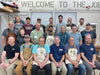 Group of people wearing aprons and casual clothing in a workshop setting with 'Welcome to The Joe' sign.